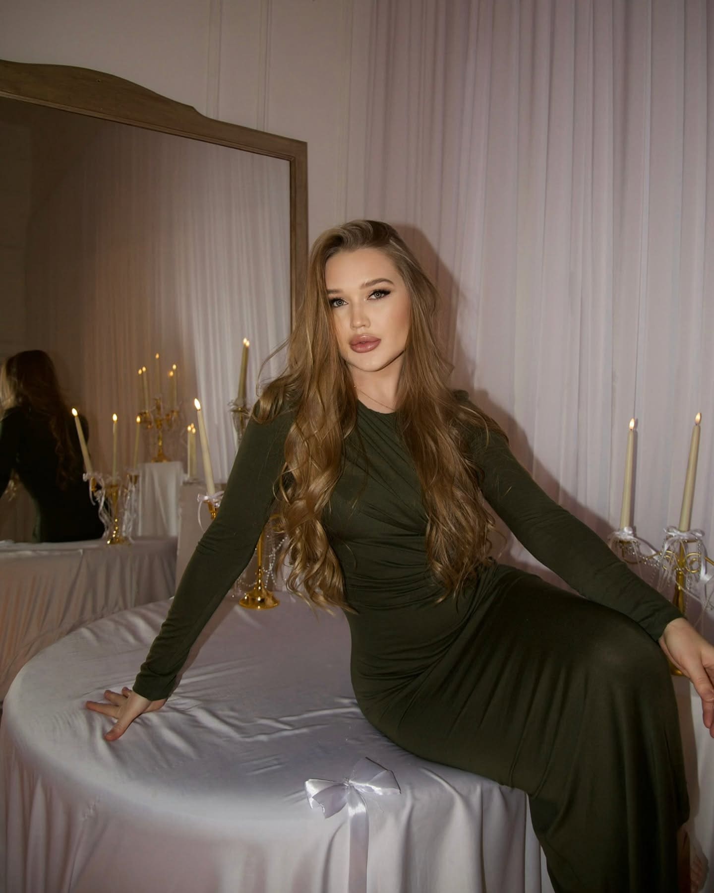 Viona sits on a white-draped table in a fitted dark green dress, with lit candles and a large mirror behind her.
