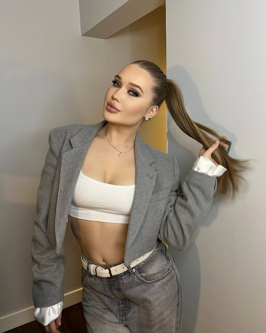 Woman in a grey blazer and jeans stands against a white wall, holding her high ponytail with one hand.