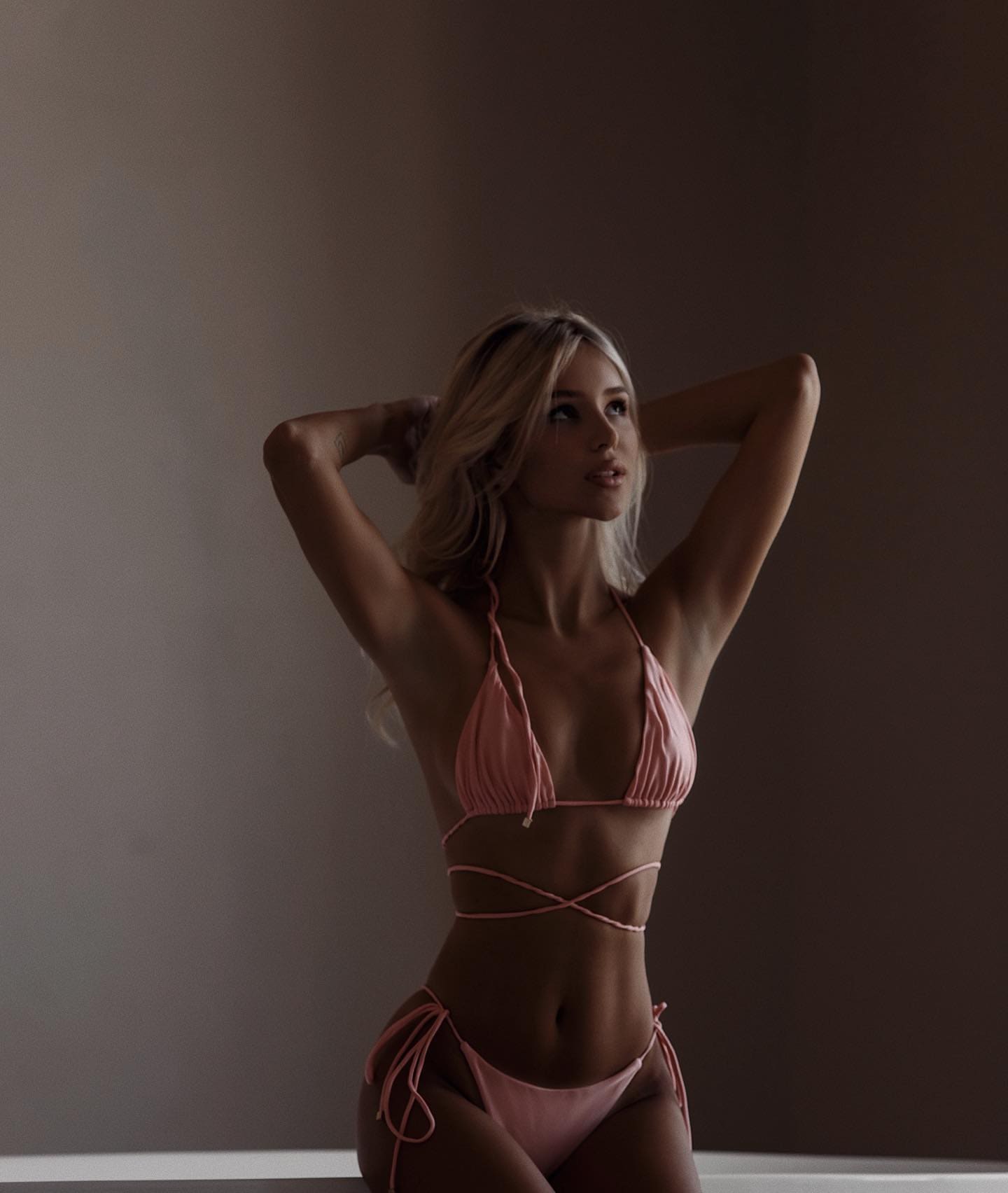 A blonde woman in a pink string bikini poses with arms raised behind her head against a dark wall.