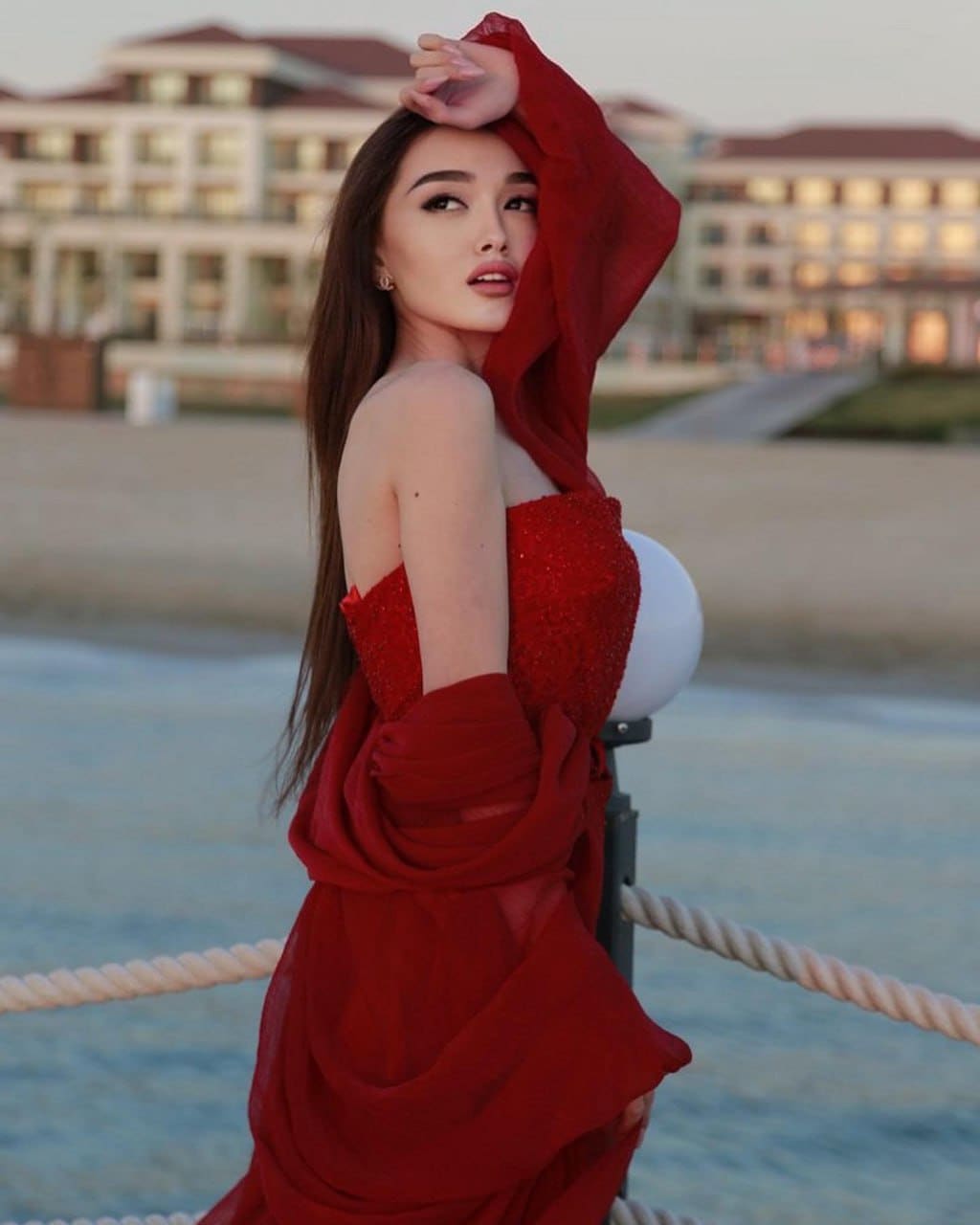 Profile view of a woman in a red sequined dress on a waterfront promenade, with water and a building in background.