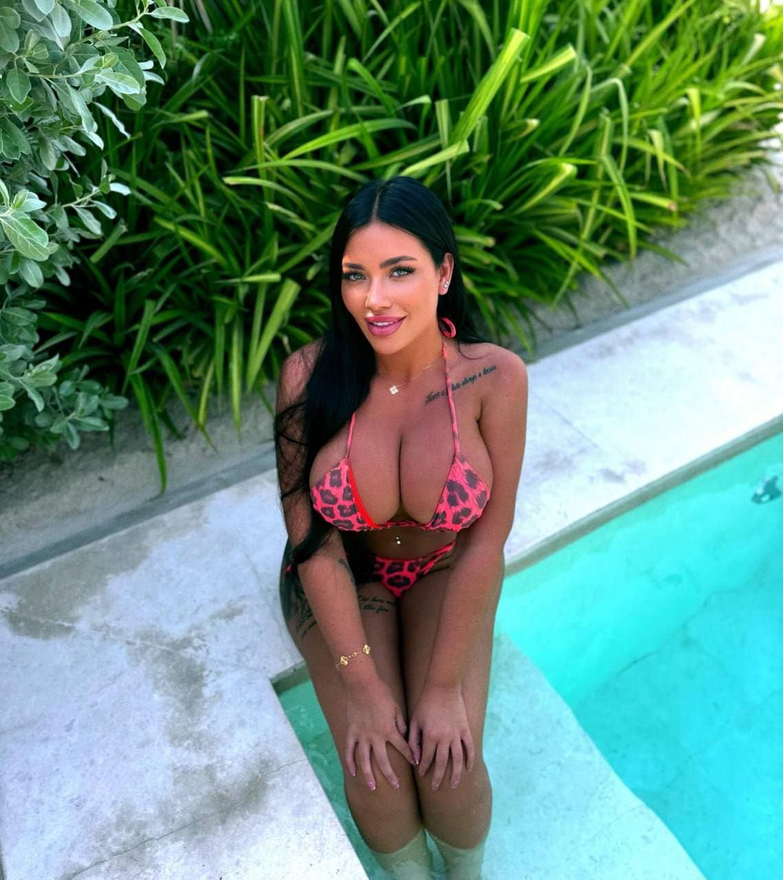 A woman sits at the edge of a turquoise pool in a pink and black leopard-print bikini, with green foliage behind her.
