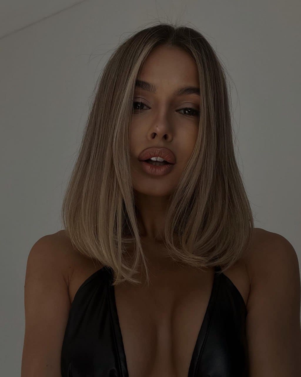 A blonde woman with a bob stands against a light wall wearing a black halter-neck top with a deep V neckline.