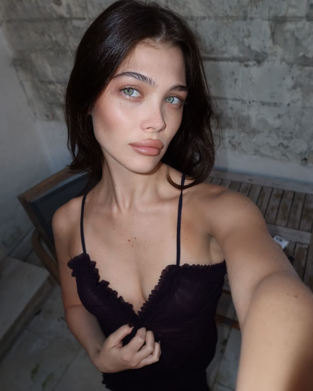 A selfie shows a woman in a black ruffled camisole standing before a weathered stone wall and a wooden bench, with shoulder-length dark hair.