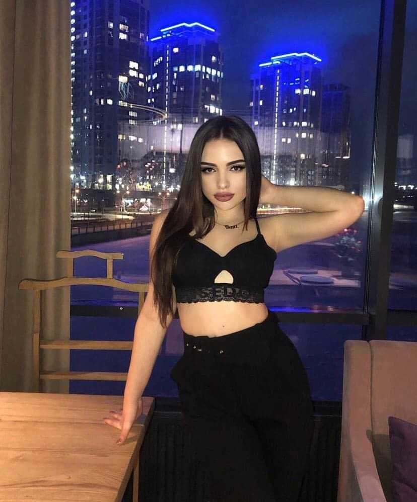 Woman in a black lace crop top and pants stands by a window showing a night skyline, one hand on a table, the other behind her head.