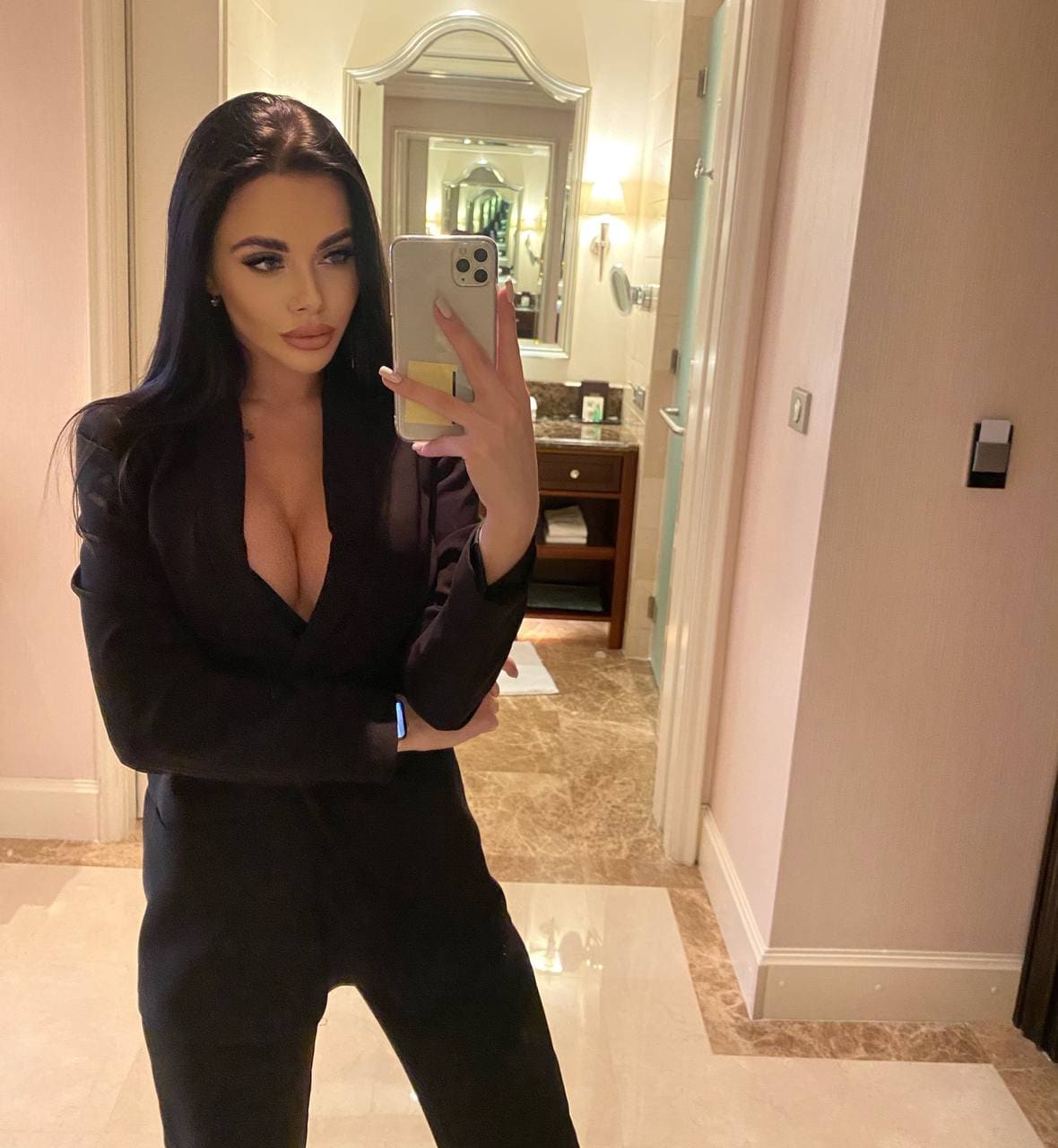 A woman takes a mirror selfie in a bathroom, wearing a black blazer with an open neckline and black trousers near a vanity.