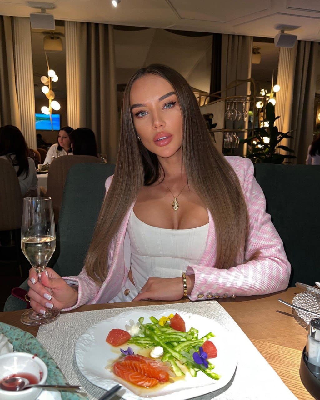 Anel sits at a restaurant table in a white top and pink blazer, holding a glass of white wine before a plate of food.