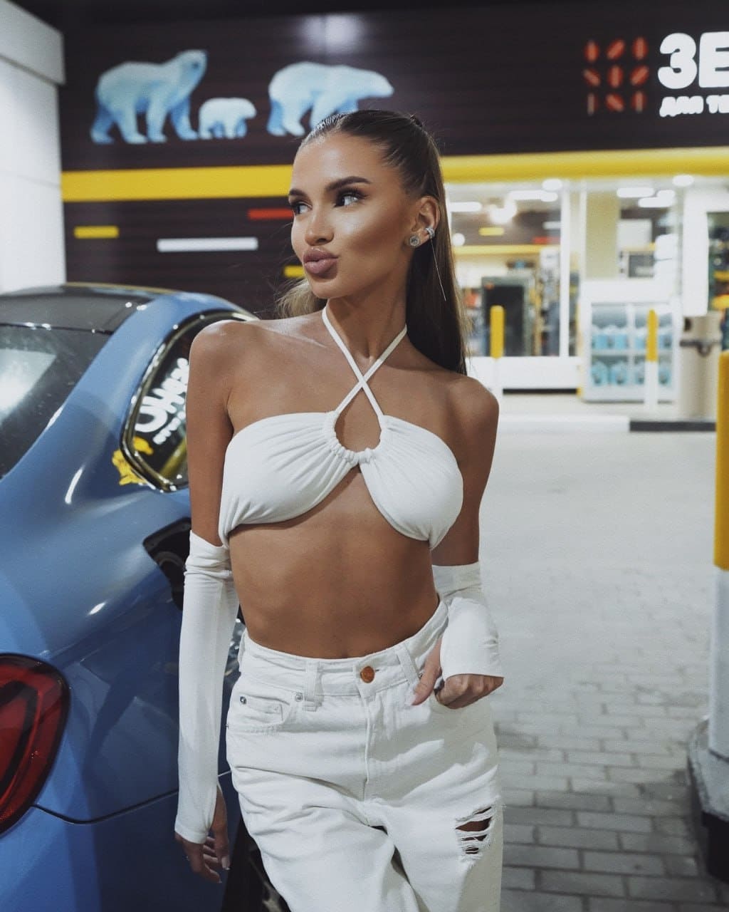 A woman with a dark high ponytail stands by a blue car in a garage, wearing a white halter top, distressed jeans, and arm sleeves.