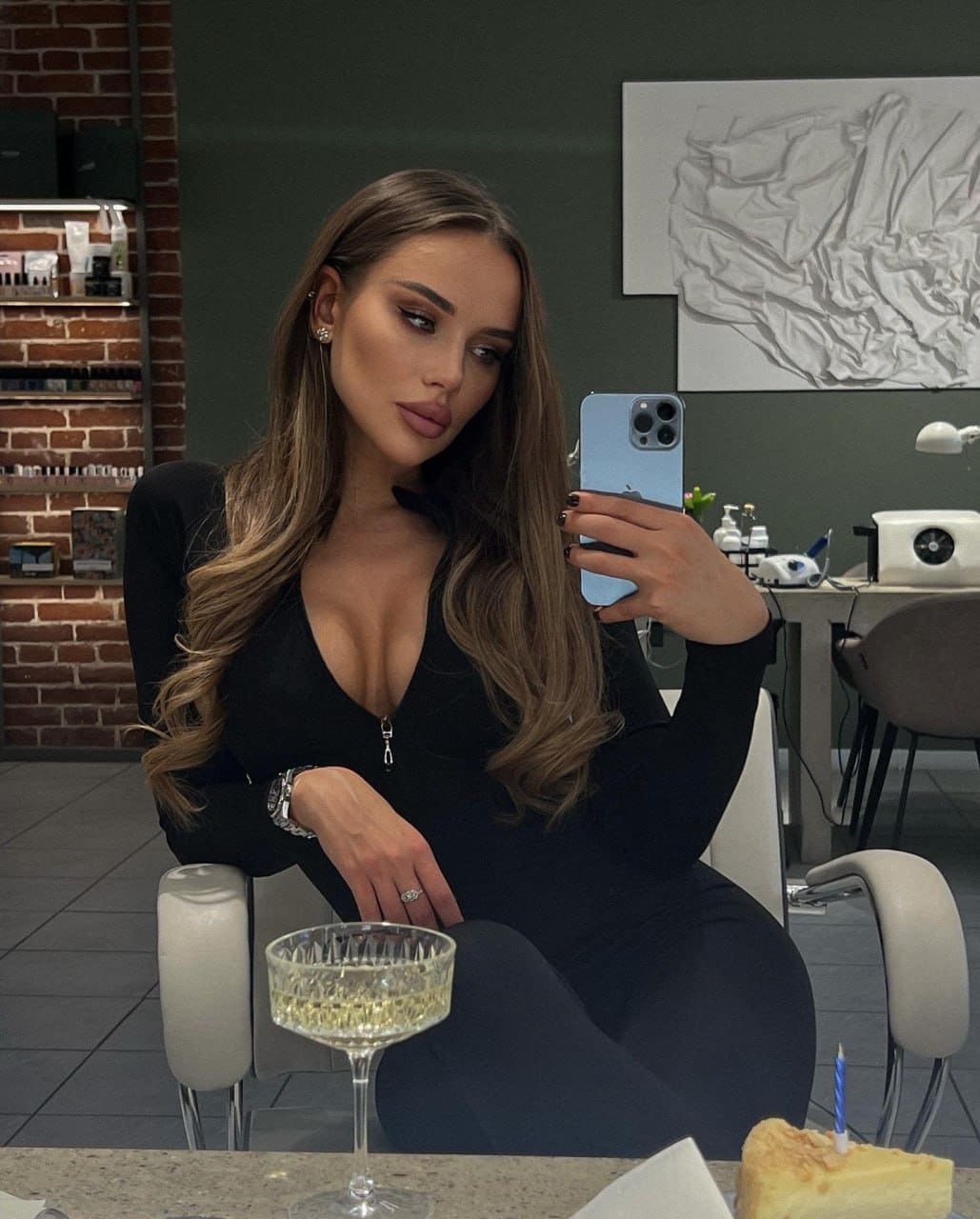 A woman takes a mirror selfie in a black V-neck dress, seated before a brick wall with champagne and cake on the table.
