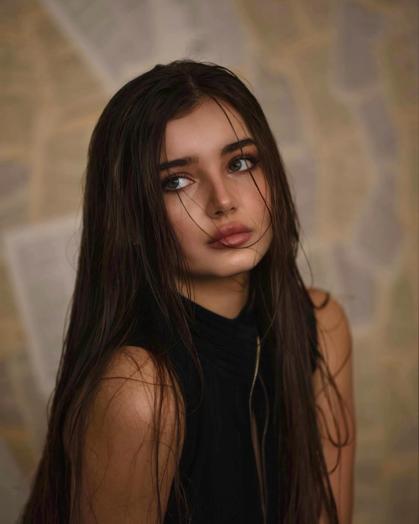 A woman with wet dark hair wears a black zippered sleeveless top, shown from the chest up against a mottled beige and grey background.