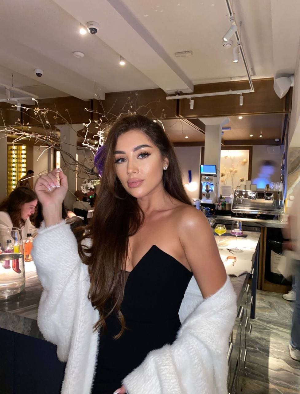 Elianora stands at a bar counter in a black strapless top and white fuzzy cardigan, one hand raised near her head.