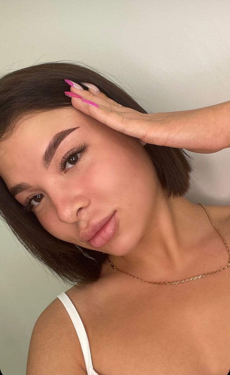 Orianna with a dark bob and pink nails wears a white spaghetti-strap top and gold chain, hand to forehead against a light wall.
