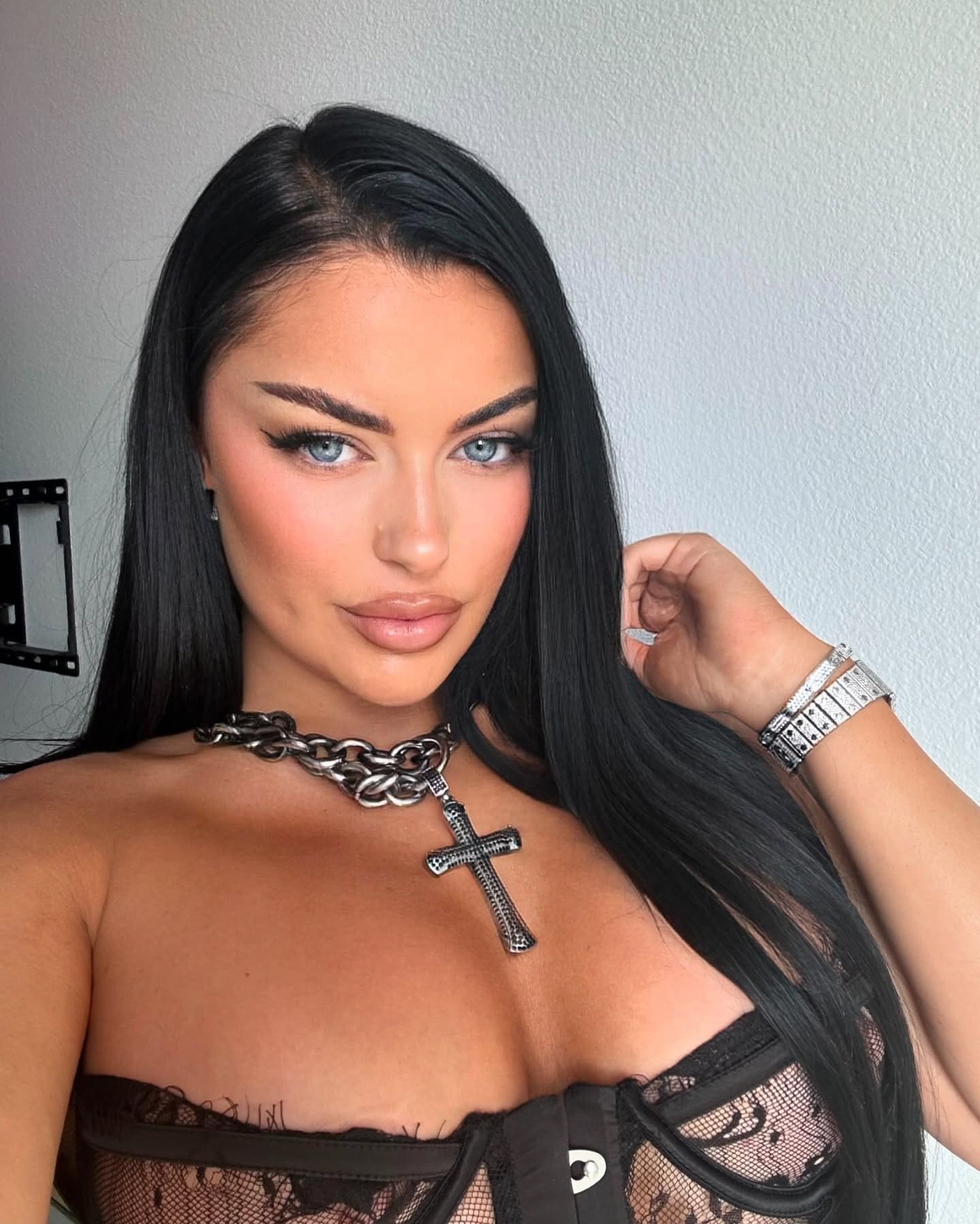 Zira wears a black lace and mesh strapless top with a cross necklace and cuff bracelet, posing against a plain light wall.