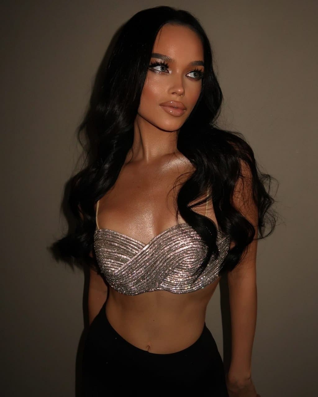 Lada stands against a brown wall wearing a silver strapless top and black bottoms, with her midriff exposed.