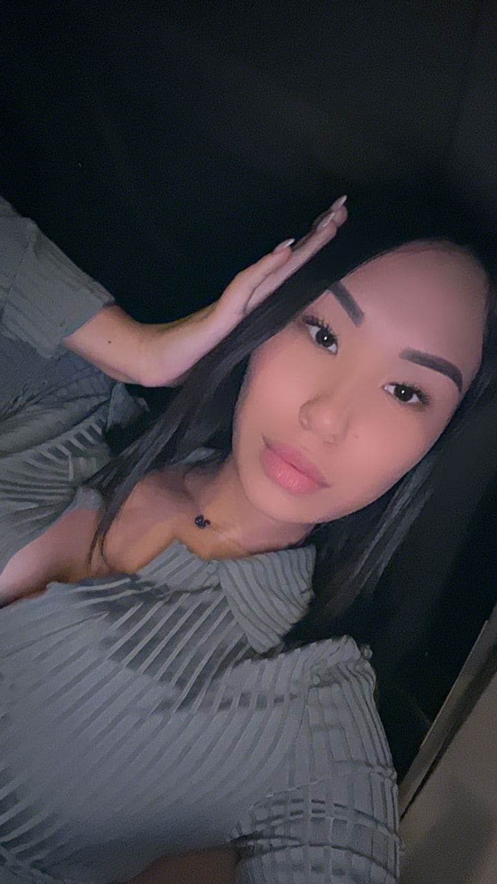 A woman rests on her side in a dark setting, wearing a grey ribbed collared top and necklace, with one hand on her forehead.