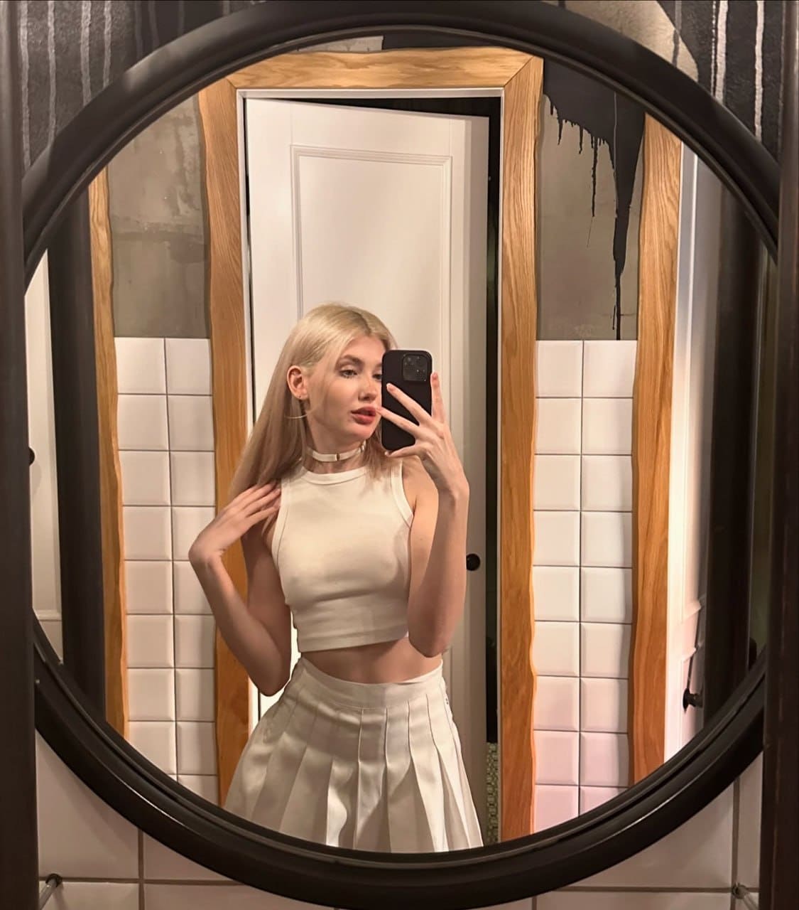 A blonde woman takes a mirror selfie in a bathroom wearing a white crop top and pleated skirt.