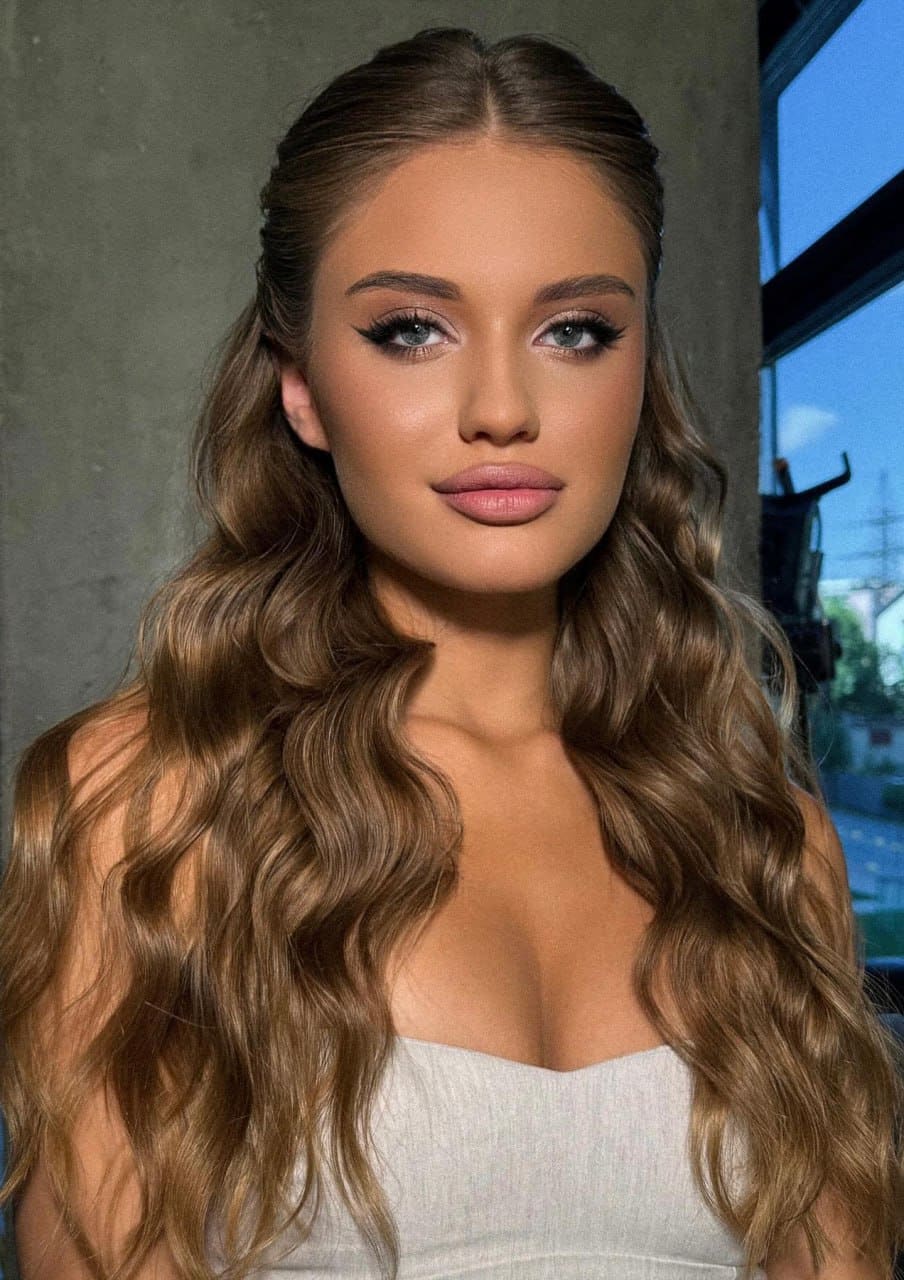 Jadora with long wavy light-brown hair faces the camera in a white strapless top, standing before a concrete wall and window.