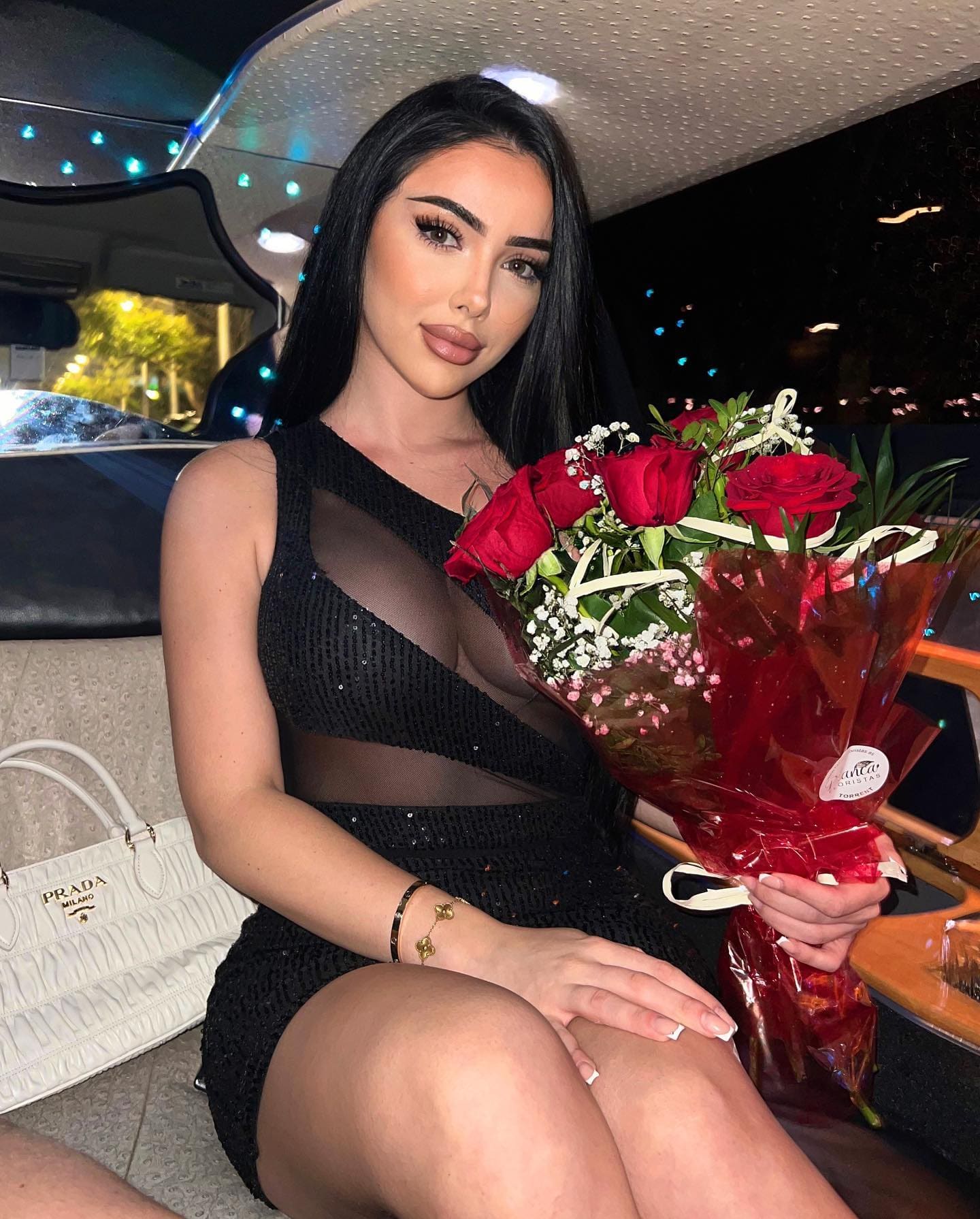 A woman sits in a car at night wearing a short black sequined dress with sheer chest cutouts, holding a bouquet of red roses with a white handbag beside her.