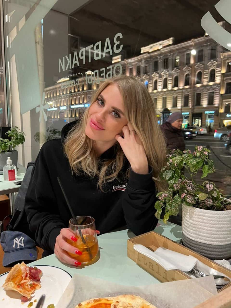 Sofil sits at a restaurant table in a black top, resting her chin on her hand beside a slice of pizza and a drink with a straw.