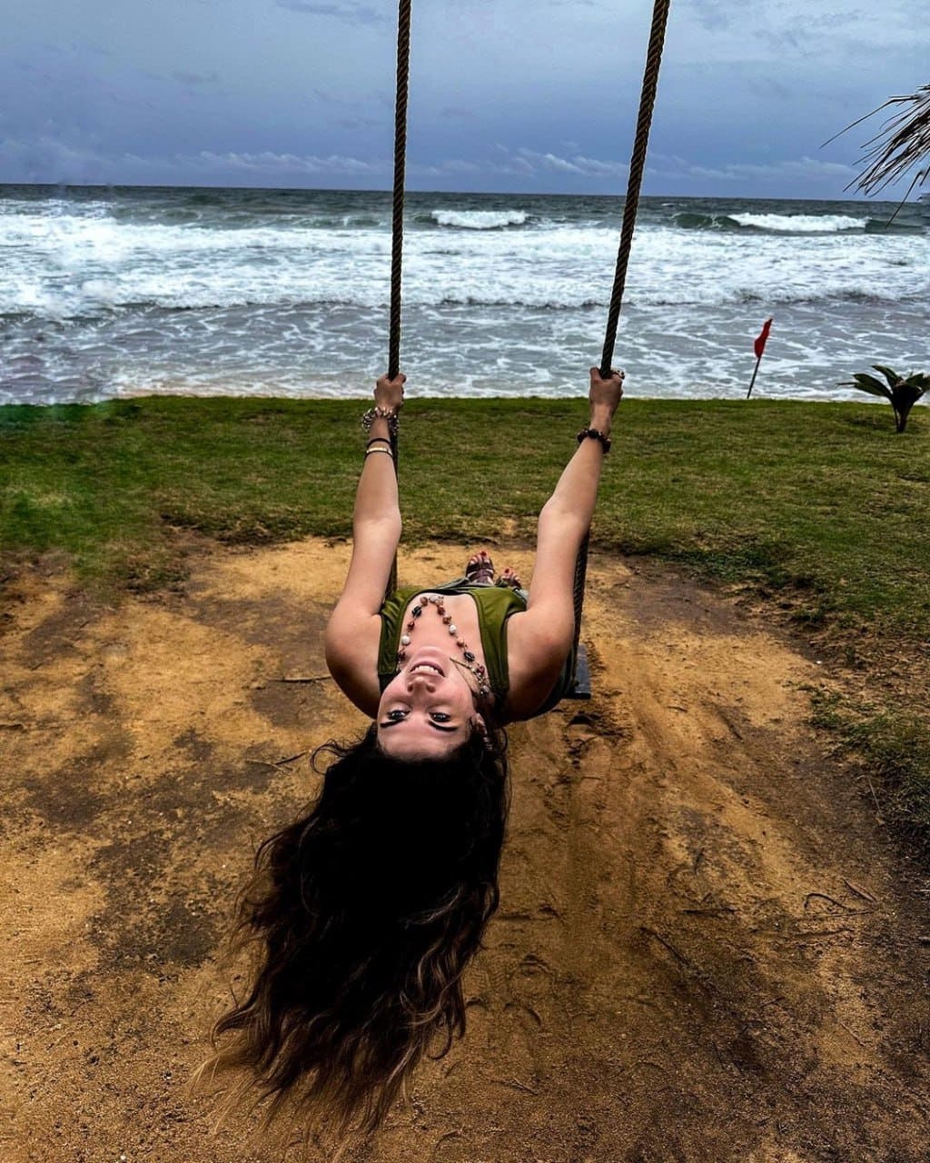 A woman hangs upside down from a rope swing on a sandy beach, wearing a green tank top and beaded necklace with ocean waves behind.