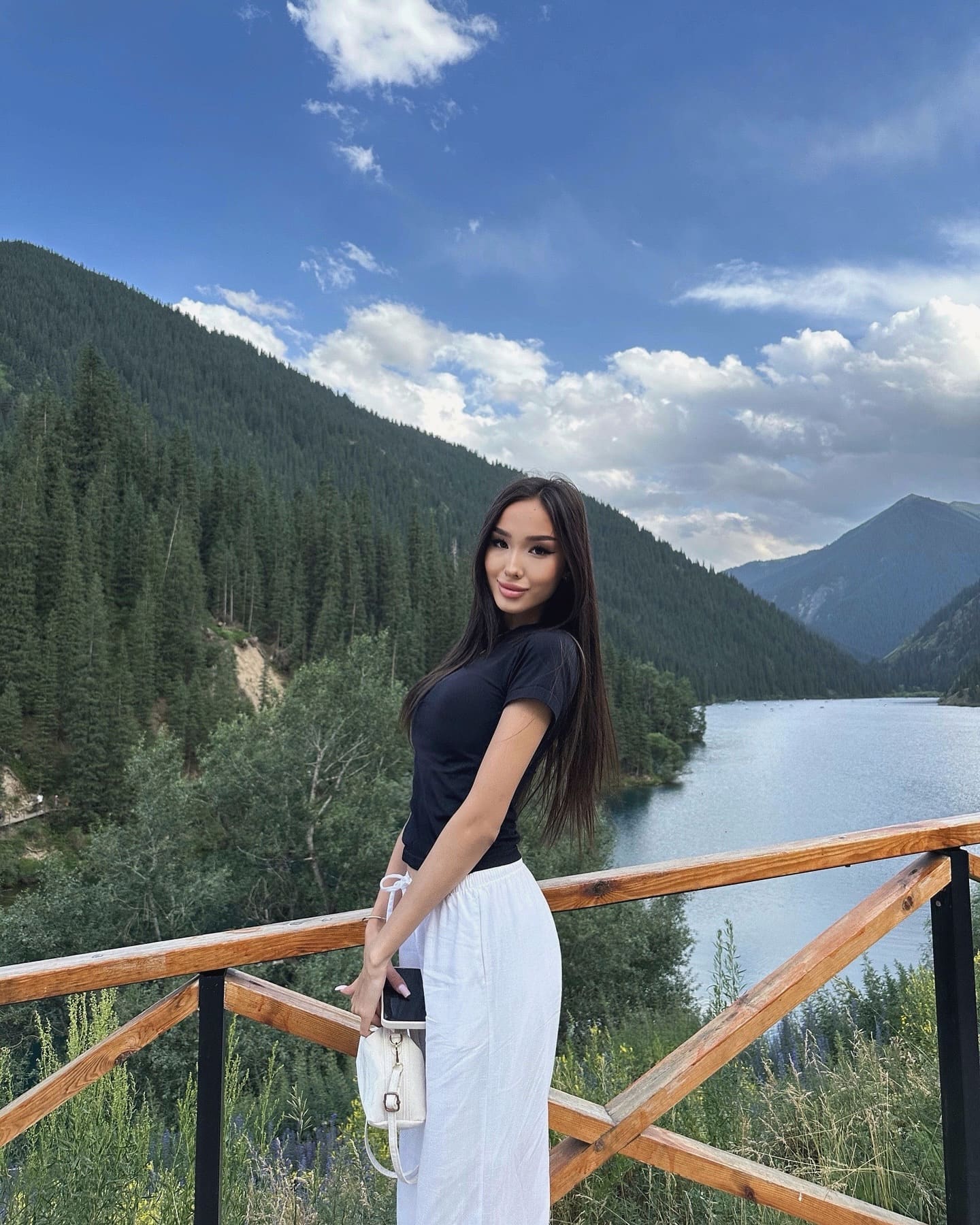 A woman stands on a wooden railing overlooking a mountain lake, wearing a black top and long white skirt while holding a small white handbag.