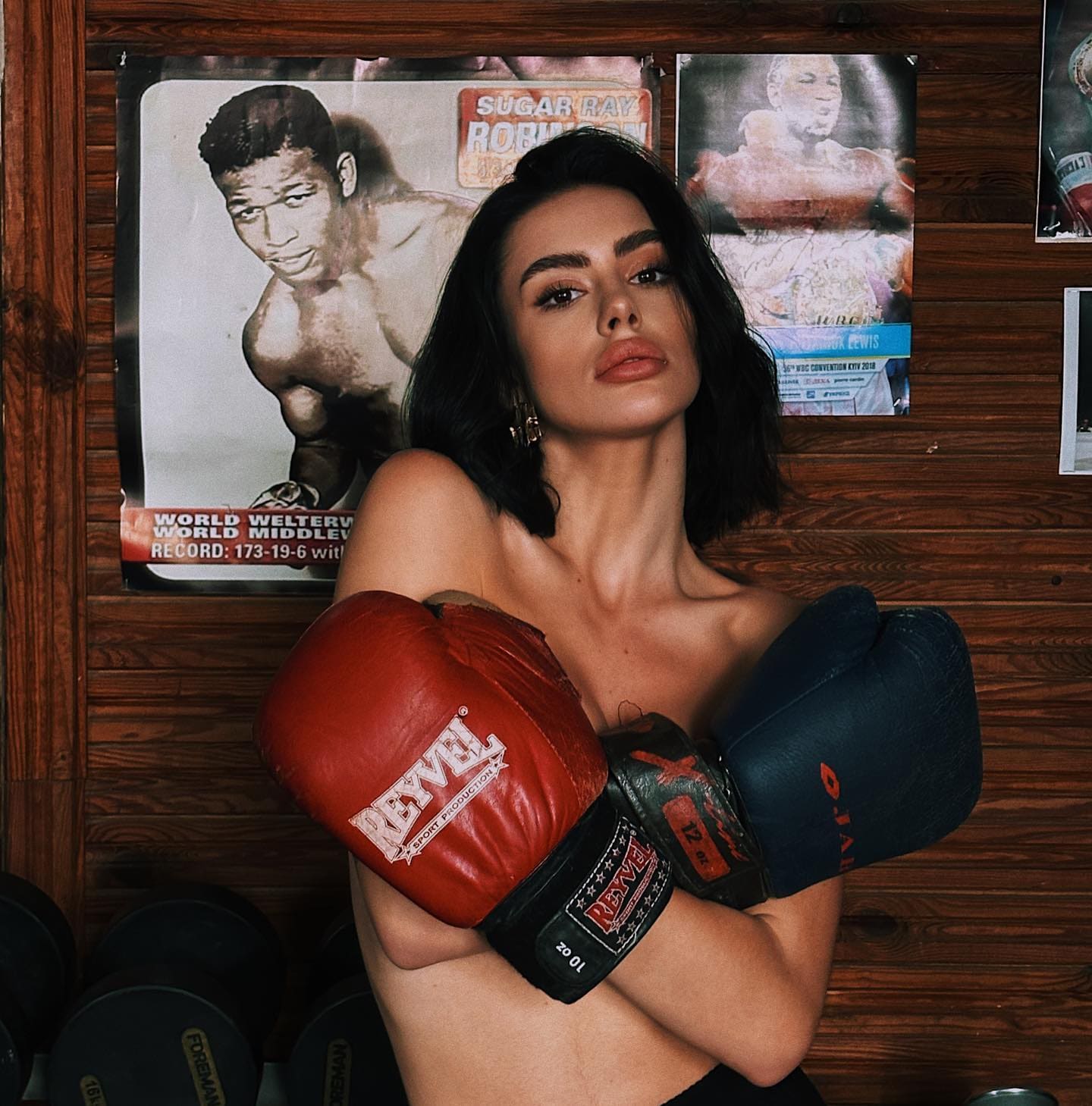 Standing with arms crossed in boxing gloves, a woman is framed by wooden paneling and posters in a gym.
