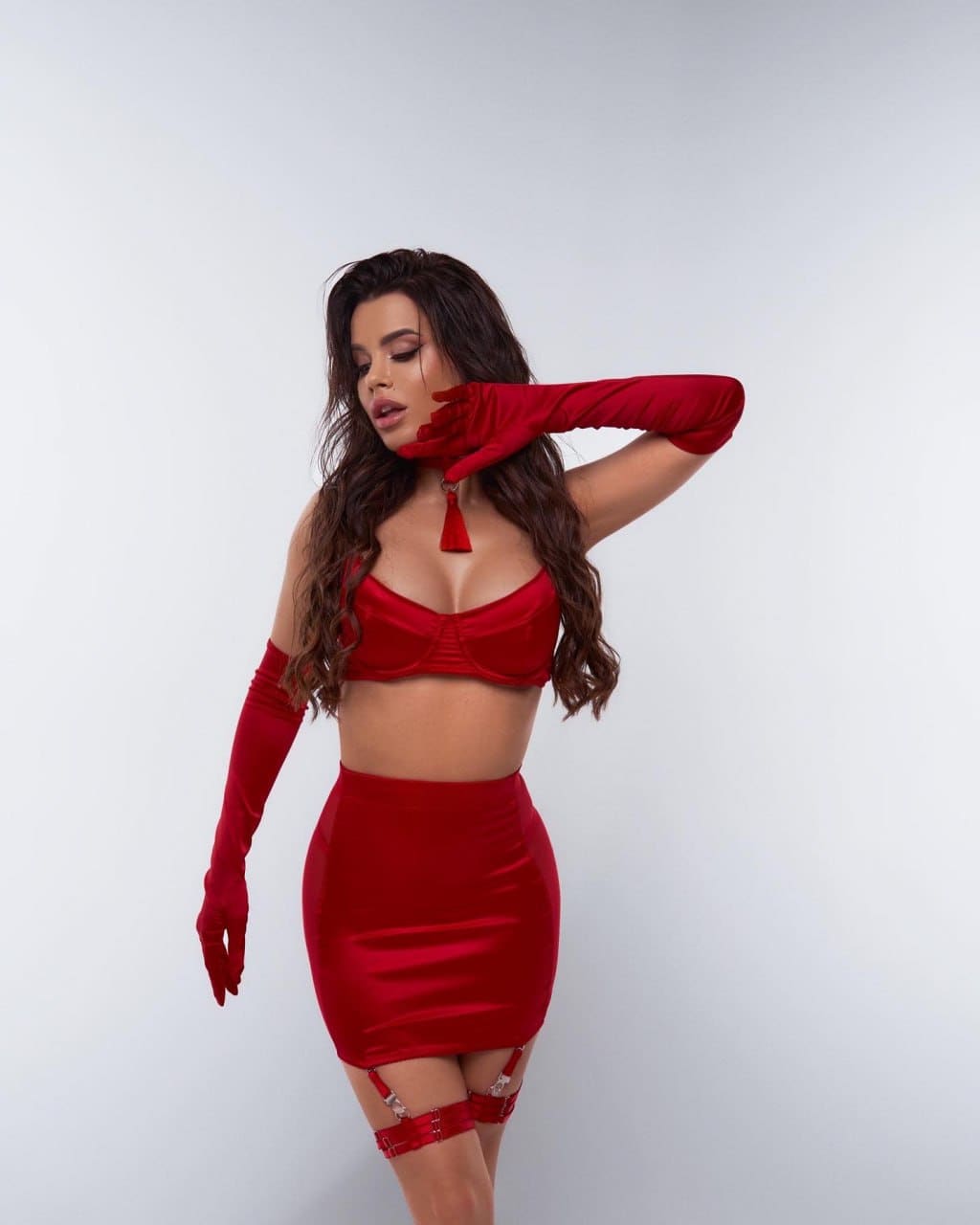 A woman in a red satin bra top, matching mini skirt, thigh-high garters, and long gloves stands against a plain light background.