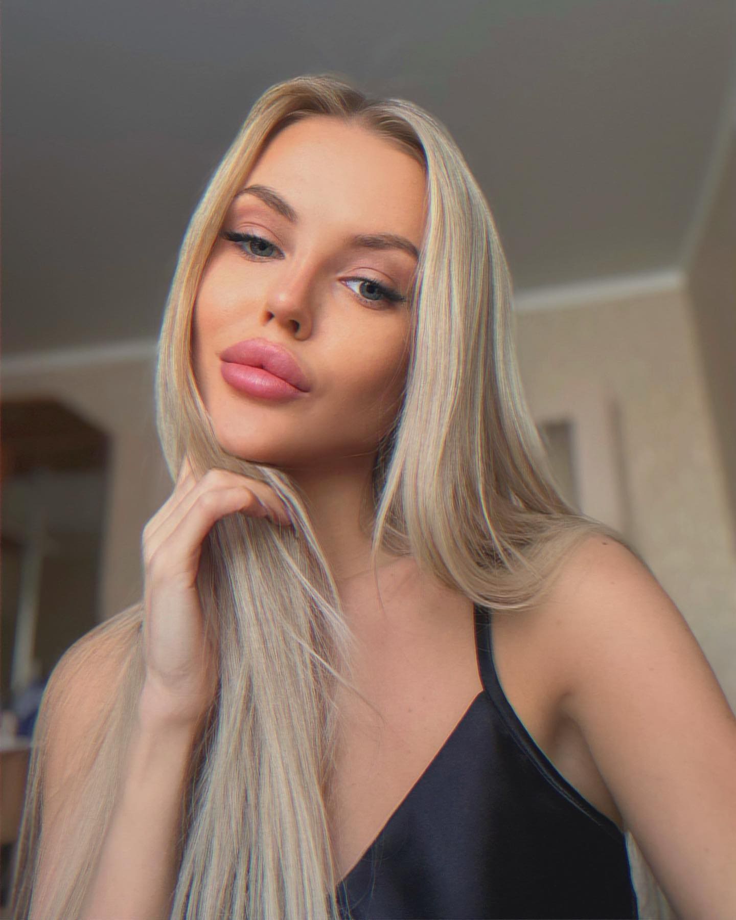 Viky stands indoors in a black sleeveless V-neck top, holding a strand of blonde hair near her chin against a blurred background.