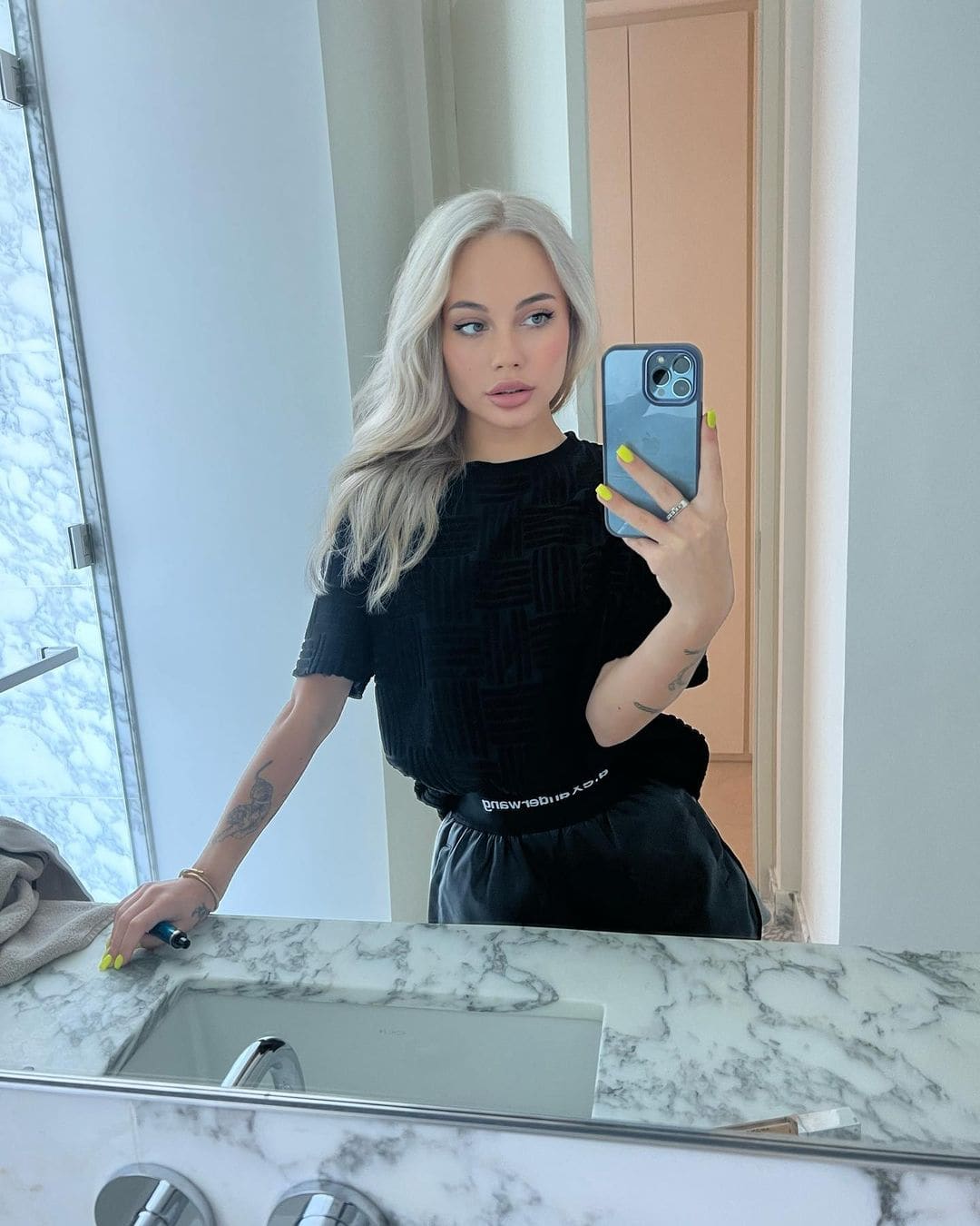 A mirror selfie shows a woman in a black textured top and pants holding a blue phone at a marble bathroom counter.