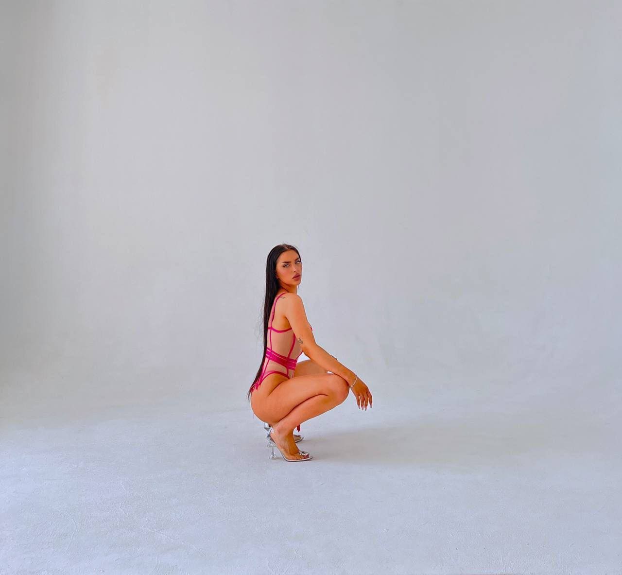 A woman in a pink strappy one-piece swimsuit crouches on a gray studio floor, looking back over her shoulder.