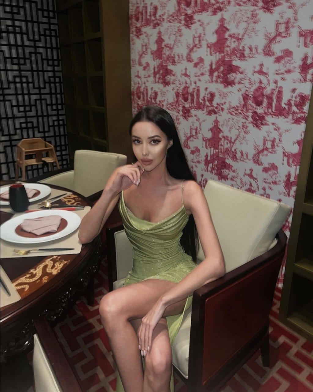 She sits at a dining table in a cream chair, wearing a short sleeveless light-green dress with a low neckline.