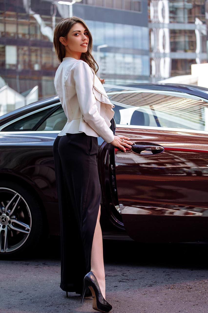 Kamilla stands in profile beside a dark car, wearing a white blouse, black high-slit trousers, and heels, with a hand on the door handle.