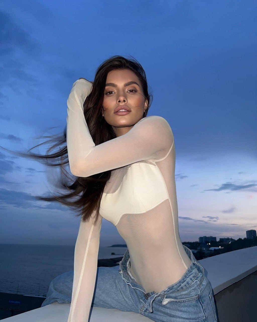 A woman sits on a rooftop ledge at dusk in a sheer top and jeans, arm raised, with the sea and city skyline in the background.