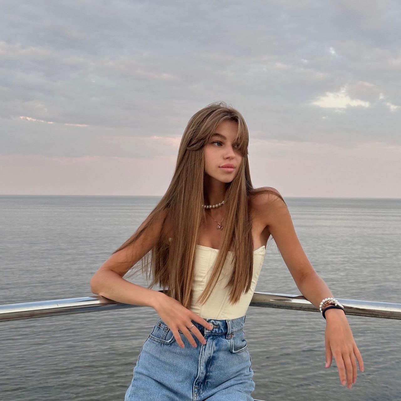 Renata stands by a railing near water in a white strapless top and light blue jeans, under a cloudy sky.