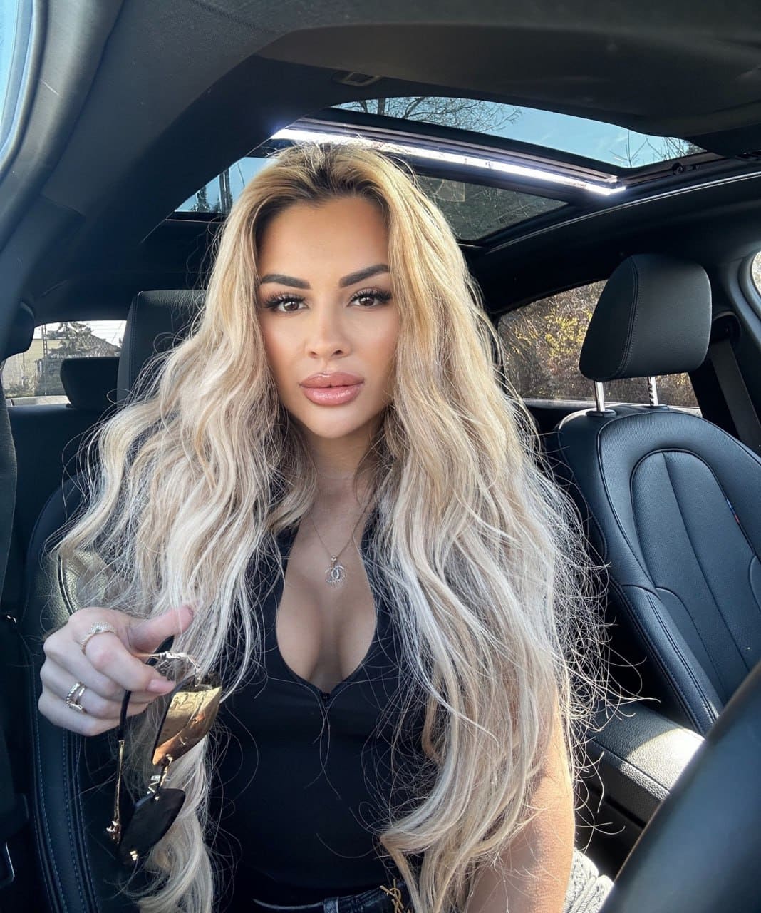 A woman with wavy blonde hair sits in a car back seat with a sunroof, wearing a black low-cut top and holding sunglasses in her hand.