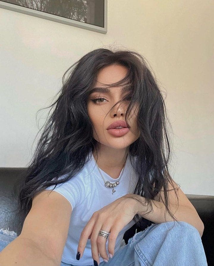 Madeline sits indoors in a light blue t-shirt and jeans, holding the camera for a selfie with a framed picture on the wall behind her.